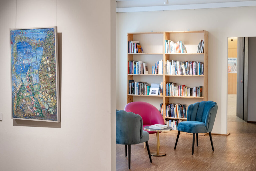 A vibrant art piece hangs on the wall beside a stylish seating area with a pink chair and blue chairs, set against a backdrop of a well-stocked bookshelf in an inviting gallery space.
